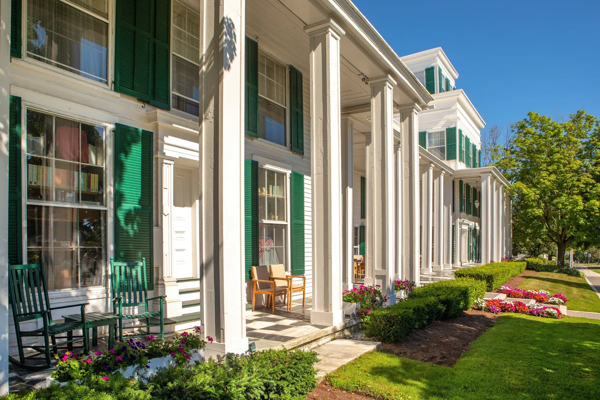 This image showcases the home's exterior, featuring green shutters, classic rocking chairs on the porch, and vibrant flower beds. The porch offers inviting spaces to relax while enjoying garden views, sunsets, or evening chats under the stars.