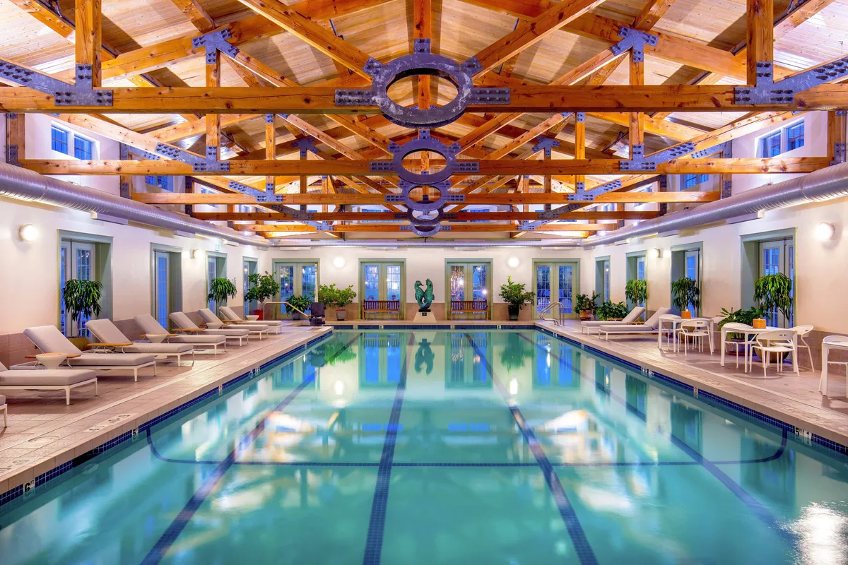 A serene indoor swimming pool featuring expansive wooden beams, stylish lounge chairs, and lush indoor plants. The atmosphere is tranquil and inviting, ideal for morning swims, poolside relaxation, or gentle exercise.