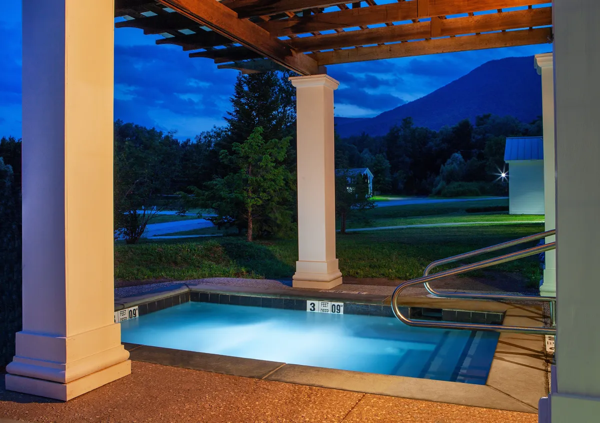 An evening exterior scene featuring an illuminated hot tub under a wooden pergola, surrounded by the natural landscape. The area is set up for socializing and is adjacent to a spot suitable for barbecues, suggesting outdoor dining and evening gatherings under the stars.