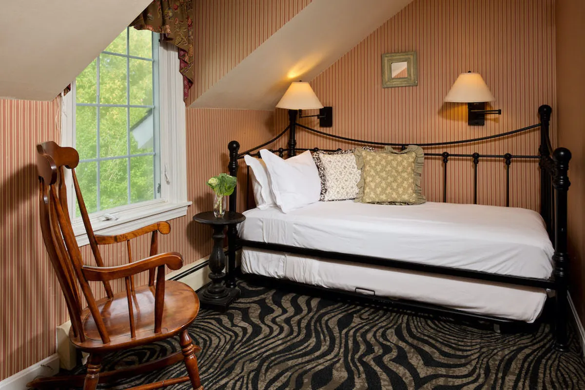 A cozy bedroom with a bed positioned near a large window, a rocking chair beside it, and sunlight streaming in, inviting a sense of relaxation. The window provides a view to the outdoors, hinting at tranquil mornings or restful afternoons, and encourages a feeling of closeness to nature.