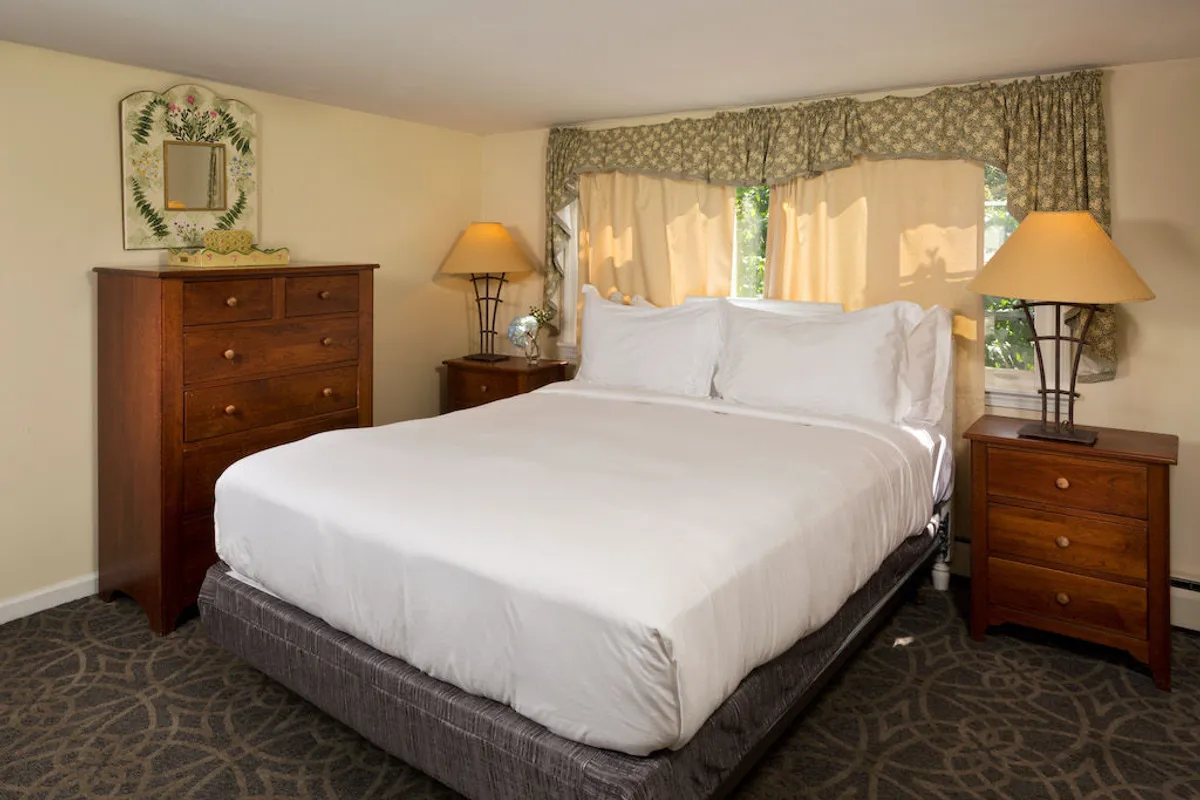 A serene bedroom featuring a queen-sized bed with soft linens, a pair of dressers, and a sunlit window framed with curtains. The décor is tranquil, with gentle colors and inviting textures that foster relaxation and comfort, ideal for adult guests or couples.