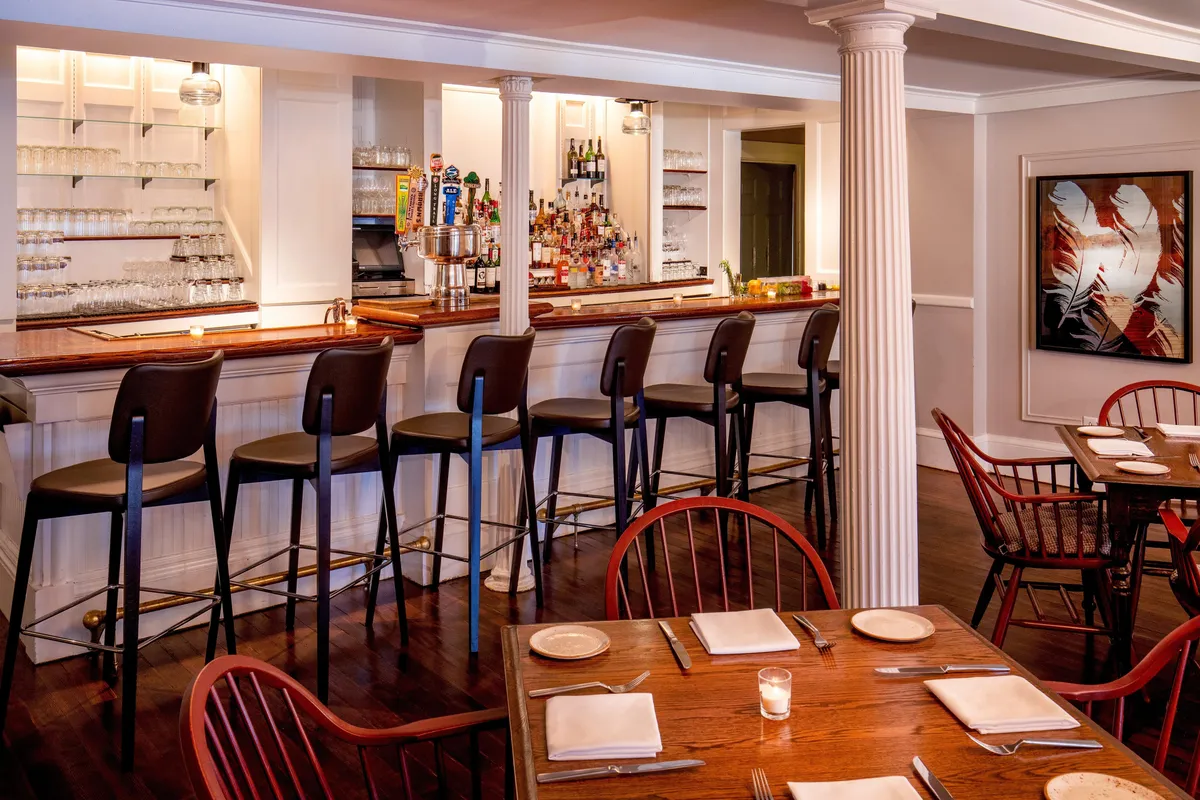 An interior scene of a welcoming bar area, featuring wooden tables for group gatherings, a bar counter, and local artwork on the walls. The space invites guests to socialize, dine, and enjoy drinks together.