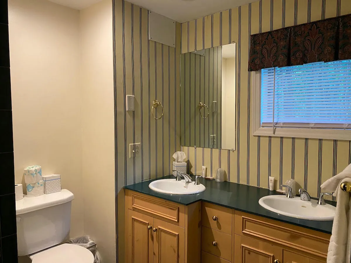 A bright bathroom with a double sink, striped wallpaper, and a window with blinds that lets in natural light. This setting suggests calm, restorative routines after spending time outside, and offers a glimpse of the house's inviting, pet-friendly atmosphere.