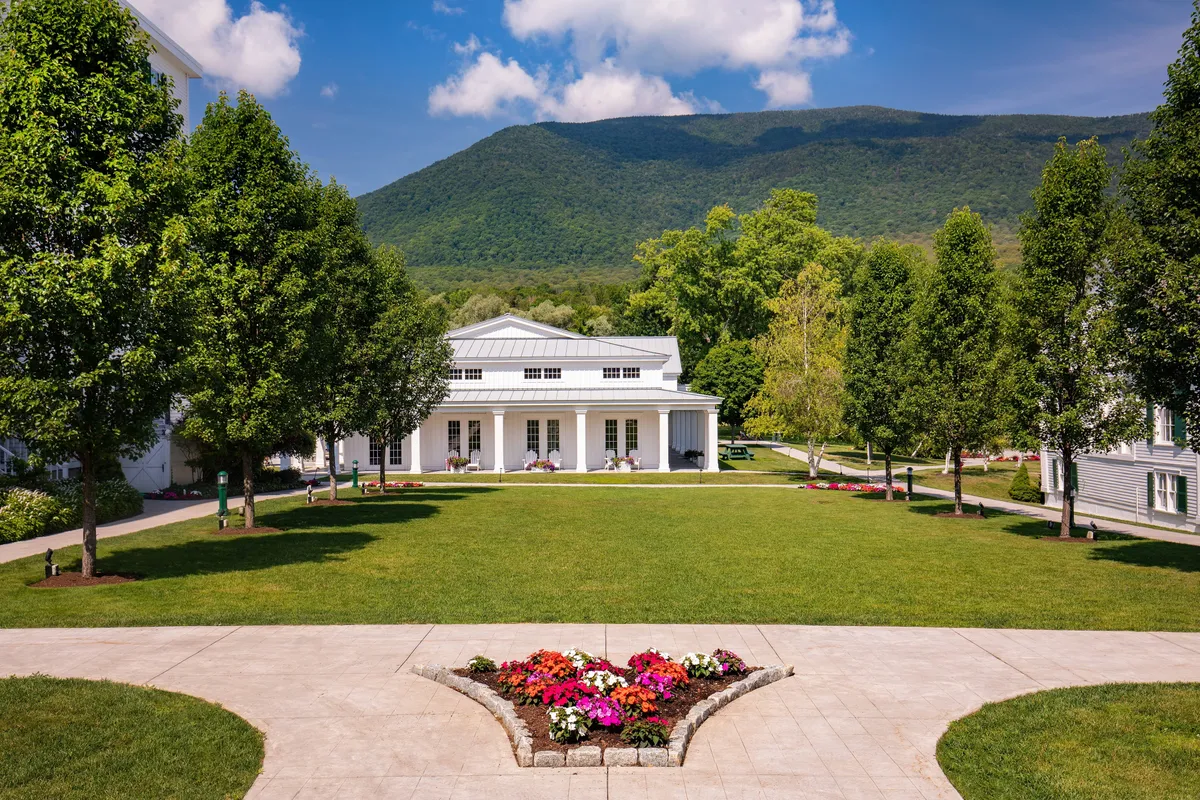 The main resort building stands framed by vibrant flower beds, a lush tree line, and a majestic mountain backdrop. The open grounds suggest ample space for family gatherings, picnics, and outdoor play.