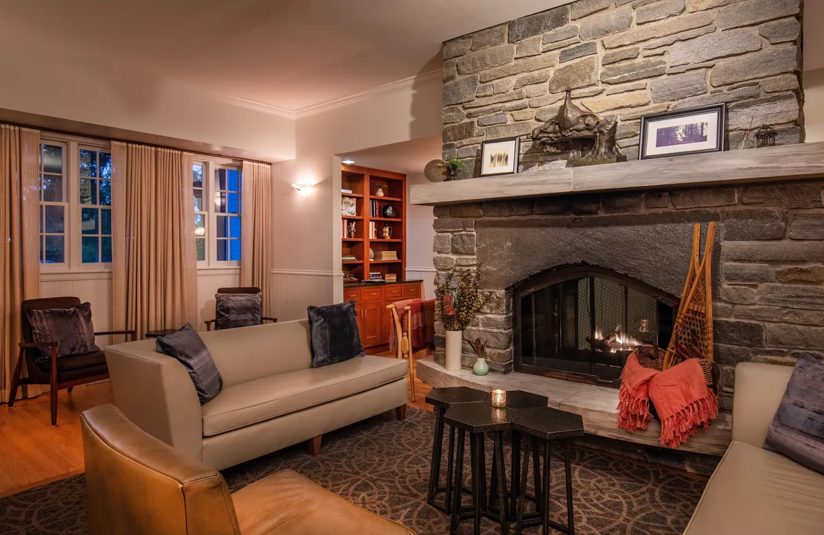 A cozy interior scene centered around a rustic stone fireplace, surrounded by comfortable seating and shelves lined with books. The inviting atmosphere suggests warmth and togetherness after a day spent outdoors.