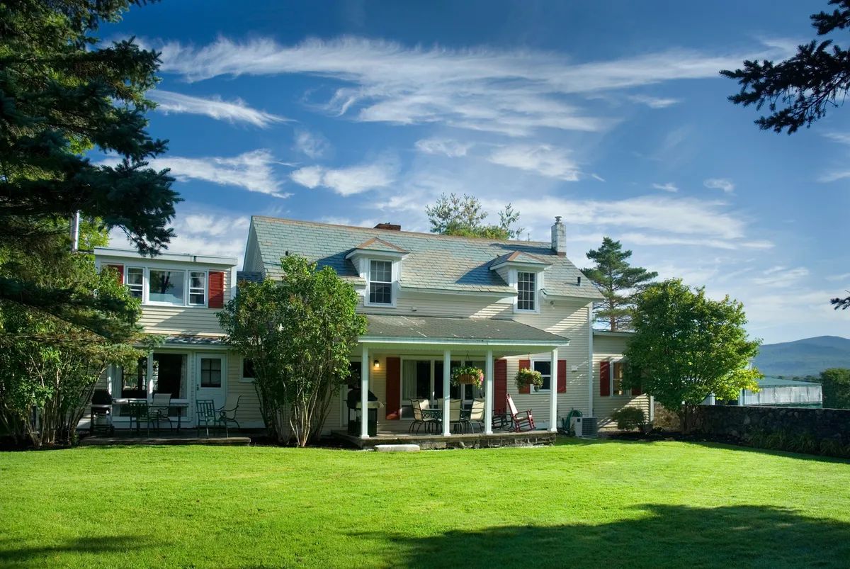 This image showcases a welcoming porch with comfortable seating, a manicured lawn, towering trees, and a view of distant mountains. The area invites guests to enjoy outdoor dining or relax while taking in the natural beauty that surrounds the home.