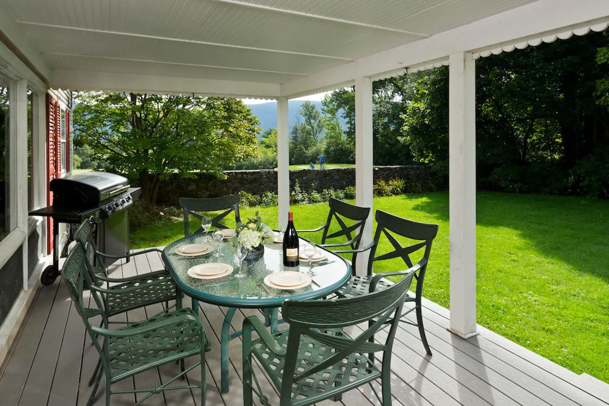 The image showcases an inviting outdoor dining area with a glass table set for a meal, a bottle of wine ready for pouring, and a grill nearby, all overlooking a lush green lawn framed by mature trees. The cheerful setting hints at sunset gatherings and festive barbecues, perfect for relaxing with friends or family in a private, scenic environment.