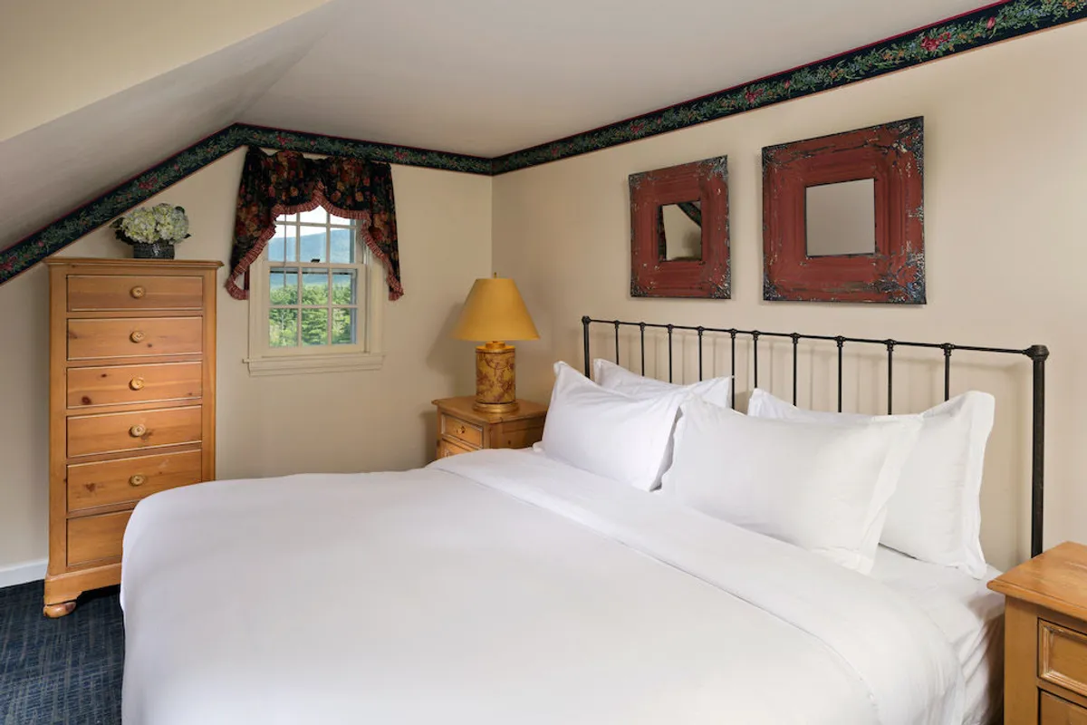 A calming bedroom setting with a bed adorned in crisp white linens, a wooden dresser, tasteful lampshade, and framed mirrors. The room exudes a sense of peace, ideal for unwinding after a day of activities, and offers flexible accommodation with its soothing ambiance.