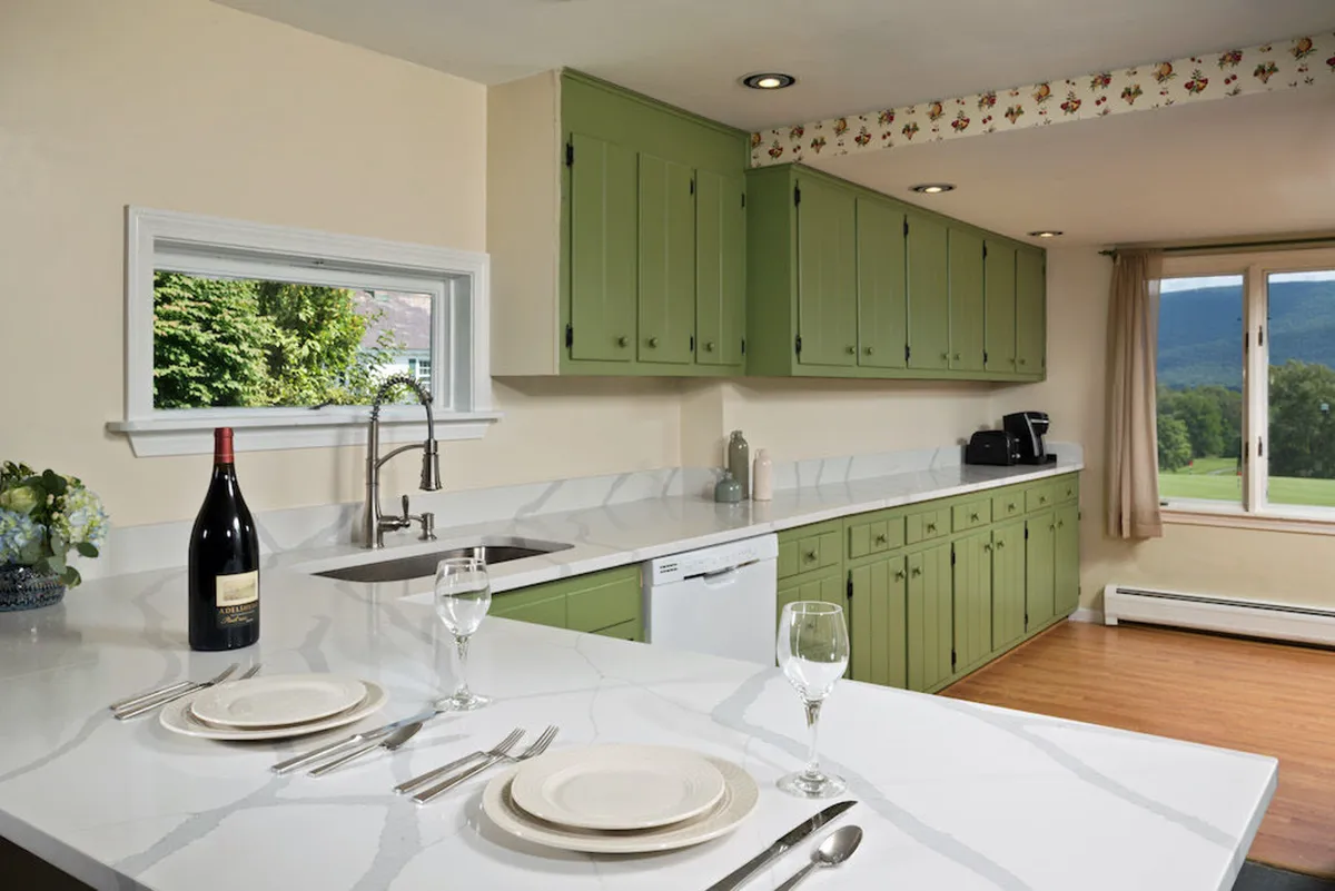A welcoming kitchen and dining area featuring green cabinetry, a marble countertop, a sunlit window, and a dining table set for a meal. A wine bottle sits on the counter, suggesting readiness for entertaining or an intimate dinner. The overall vibe is cozy and inviting, encouraging gatherings and shared meals.
